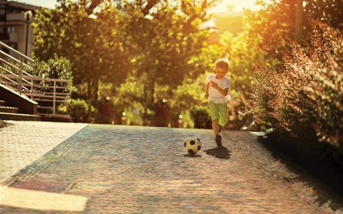 Little boy is playing football in the city. The boy aged 6 is running after the football and smiling. Sunny summer day in city residential area.