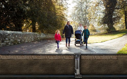 A photo with two women and three children, one in a stroller, walking in the park. The Smart Probe system is shown below the asphalt.