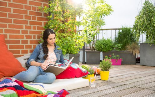 Woman surrounded by colorful cushions and blankets on rooftop terrace flipping through photo album in front of brick wall