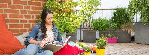 Woman surrounded by colorful cushions and blankets on rooftop terrace flipping through photo album in front of brick wall
